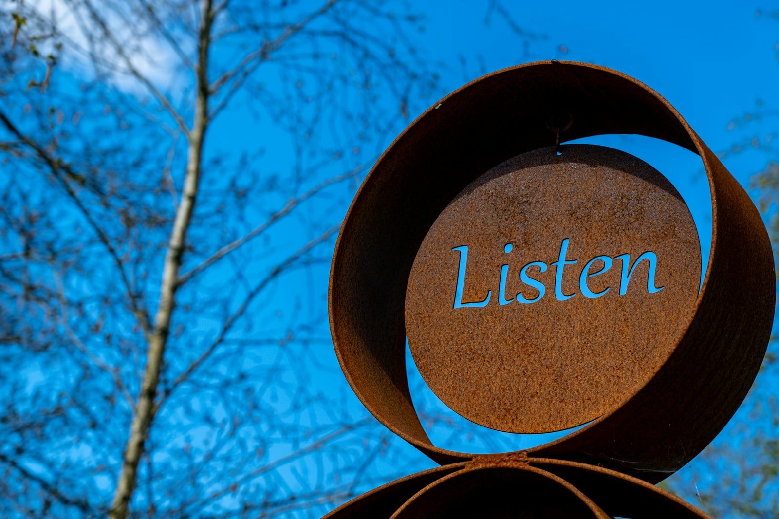 A weathered metal sculpture cut to read 'Listen' in cursive script, set against a clear blue sky and bare branches.