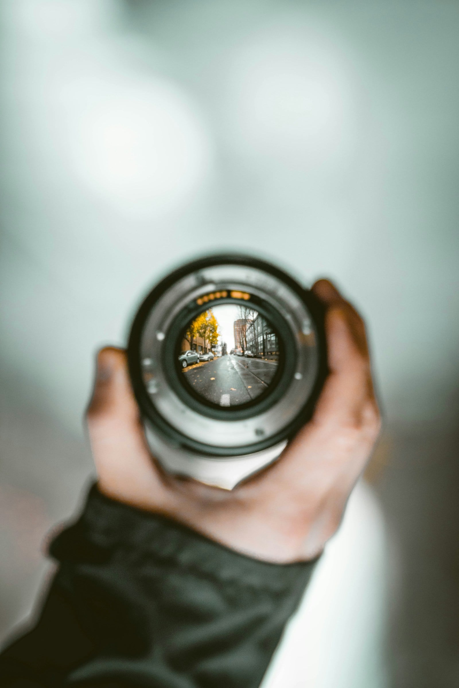 A hand holds a detached camera lens up at arm's length, framing a sharp view of a city street through the lens while everything around it falls into soft blur.