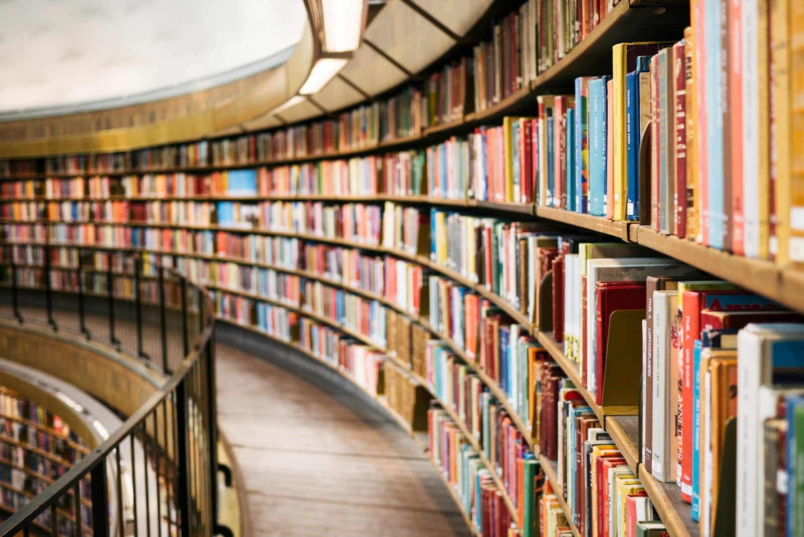 A long, curved corridor of bookshelves filled with brightly colored book spines, lit by warm overhead lights, viewed from a low angle along the curve.