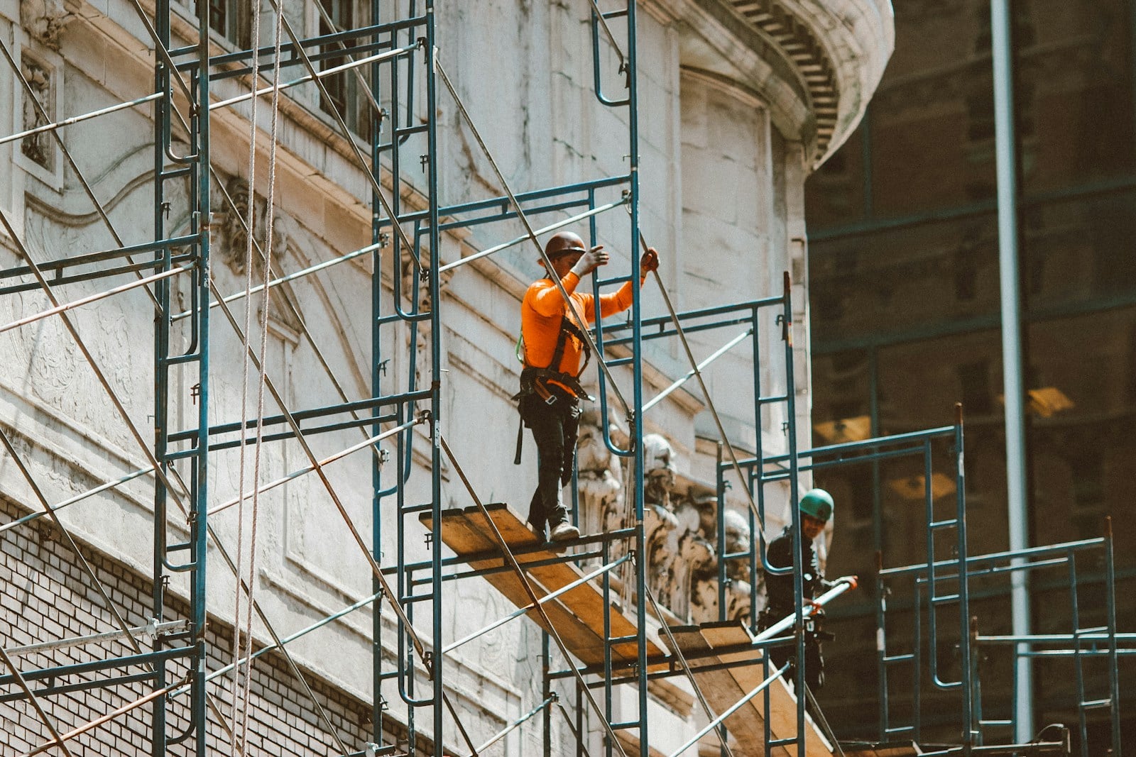 Construction workers in safety harnesses stand on metal scaffolding against the side of an ornate building, working at height.