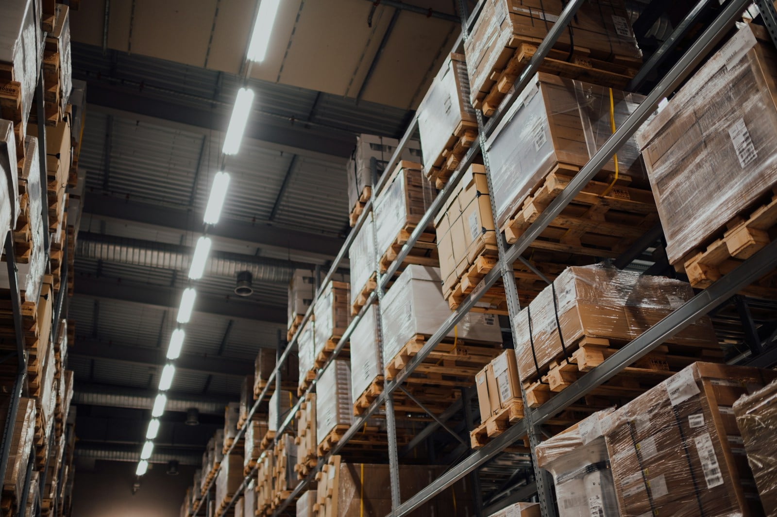 Tall industrial warehouse racks stacked with cardboard boxes on wooden pallets, fluorescent ceiling lights stretching into the distance.