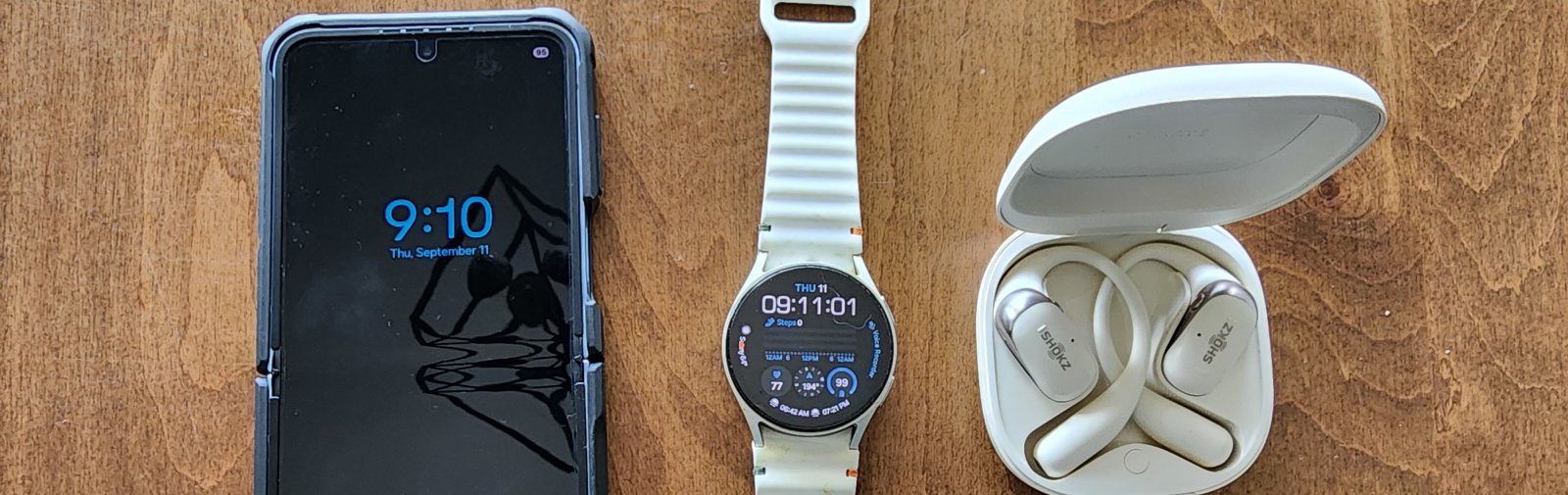 Samsung Galaxy Z Flip 6 phone, Galaxy Watch 7, and SHOKZ OpenFit 2 earbuds arranged on a wooden table, used for timers, alarms, and 45/15 breaks.
